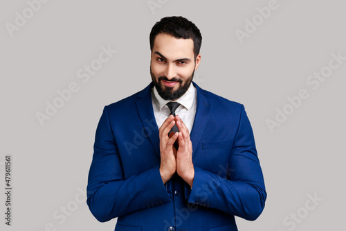 Cunning bearded man clasping hands and planning evil tricky prank or scheming, cheating with sly smile, wearing official style suit. Indoor studio shot isolated on gray background. photo