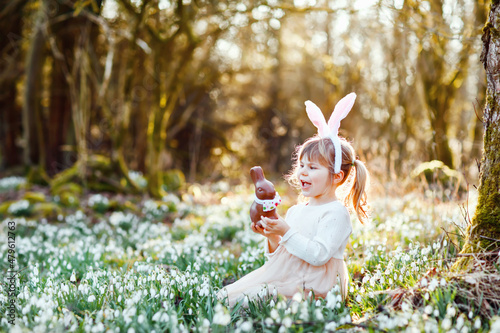 Fototapeta Naklejka Na Ścianę i Meble -  Adorable little girl with Easter bunny ears eating chocholate figure in spring forest on sunny day, outdoors. Cute happy child with lots of snowdrop flowers. Springtime, christian holiday concept.