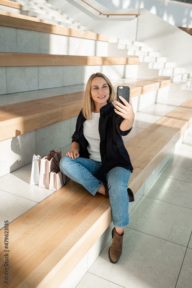 Stylish blonde girl sitting in a mall with purchases using a mobile phone to take a selfie Stylish blonde girl sitting in a mall with purchases using a mobile phone to take a selfie