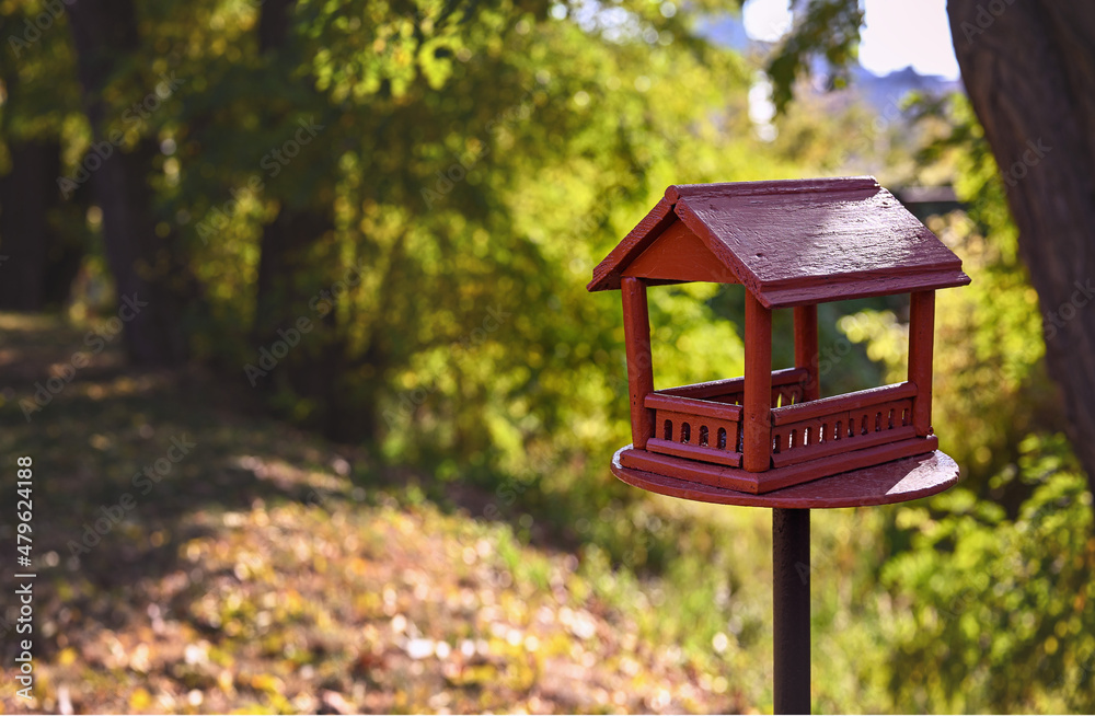 Red wooden bird feeder (birdhouse) with a roof in a city park. Sunny day, autumn. The feeder stands on a metal stand on the ground. Free space for text.