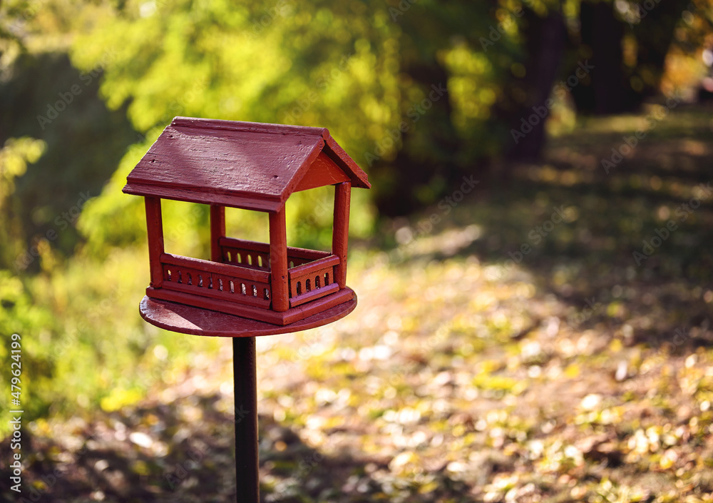 Red wooden bird feeder (birdhouse) with a roof in a city park. Sunny day, autumn. The feeder stands on a metal stand on the ground. Free space for text. Red wooden bird feeder (birdhouse) with a roof in a city park. Sunny day, autumn. The feeder stands on a metal stand on the ground. Free space for text.