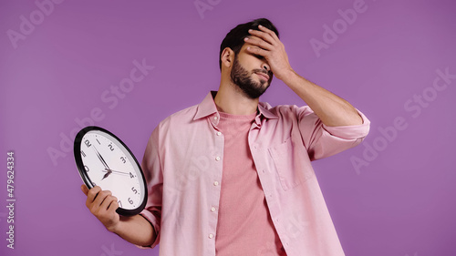 worried young man holding clock and showing face palm isolated on purple