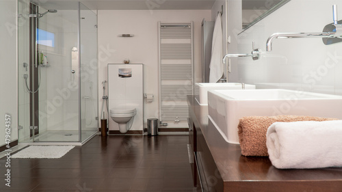 Modern bathroom in white and brown with two sinks, toilet, shower and wooden furniture