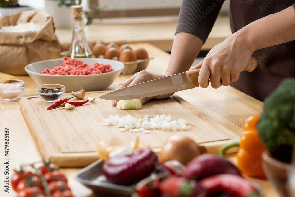 Hands of young female chopping fresh onion on wooden board while preparing italian pasta with vegetables and minced meat