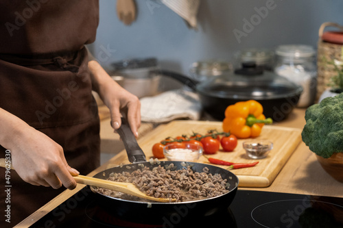 Hands of woman in apron holding wooden spoon while standing by electric stove and mixing minced meat in frying pan