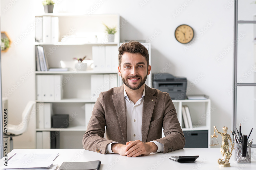 Happy young lawyer in formalwear sitting by desk in office and looking at camera on background of shelves with folders Happy young lawyer in formalwear sitting by desk in office and looking at camera on background of shelves with folders