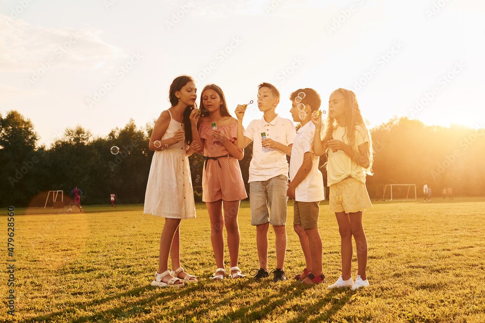 Illuminated by sunlight. Standing on the sportive field. Group of happy kids is outdoors at daytime Illuminated by sunlight. Standing on the sportive field. Group of happy kids is outdoors at daytime