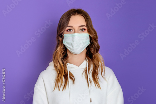Young woman wearing a medical mask and white hoodie stands isolated on a a lilac background in the studio