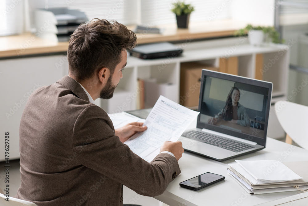 Young businessman with paper communicating with female on laptop screen during online meeting while sitting by desk in office Young businessman with paper communicating with female on laptop screen during online meeting while sitting by desk in office