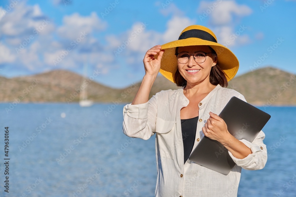 Portrait of happy mature woman in straw hat with laptop on the beach, copy space