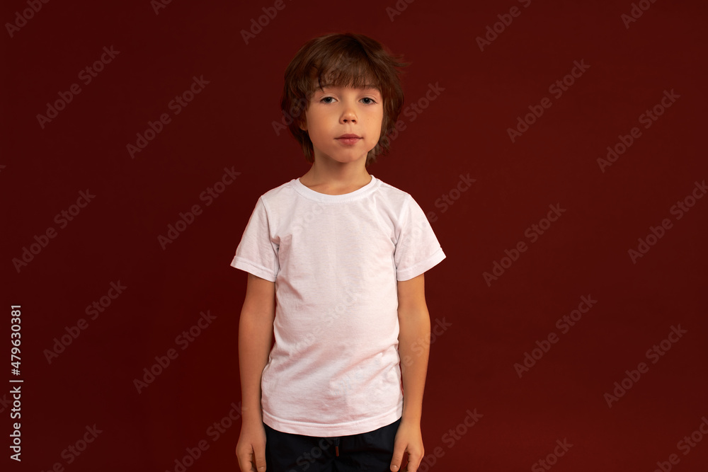Portrait of cute Caucasian 6-years-old child standing against red studio wall with calm face expression, looking at camera dressed in white t-shirt with copy space for your text or content Portrait of cute Caucasian 6-years-old child standing against red studio wall with calm face expression, looking at camera dressed in white t-shirt with copy space for your text or content