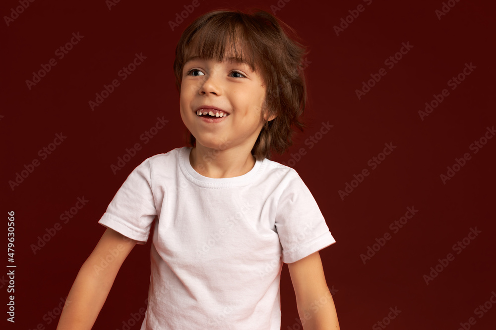 Close-up of funny-looking 6-7 years-old brunette boy in white sport shirt smiling showing his new upper permanent teeth, looking aside with cute laughing face. Lovely kids. Happy childhood Close-up of funny-looking 6-7 years-old brunette boy in white sport shirt smiling showing his new upper permanent teeth, looking aside with cute laughing face. Lovely kids. Happy childhood