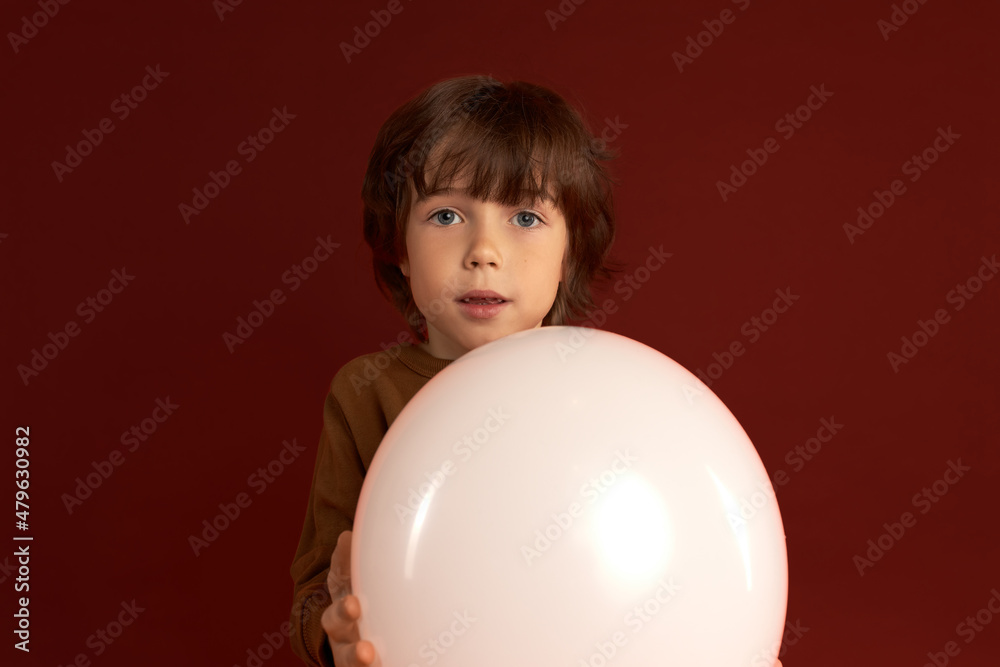 Cute shaggy perplexed Caucasian kid of 6 standing against red background in knitted brown jumper holding white ball with copy space for your text in hands, having thoughtful face expression Cute shaggy perplexed Caucasian kid of 6 standing against red background in knitted brown jumper holding white ball with copy space for your text in hands, having thoughtful face expression
