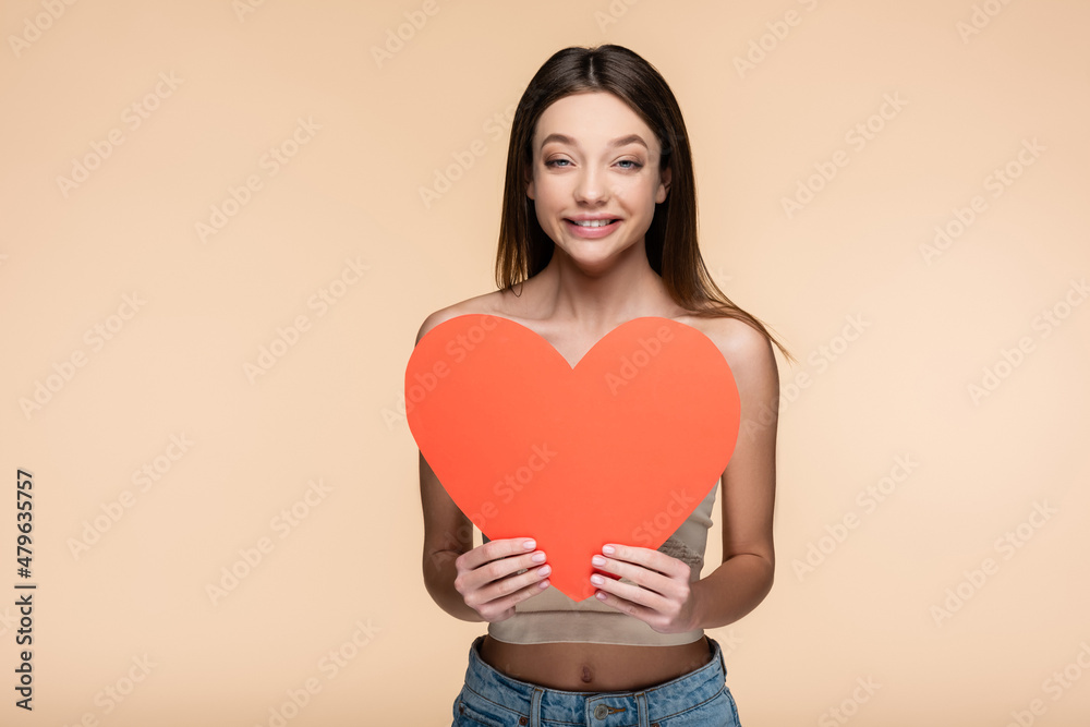 positive young woman in crop top holding red paper heart isolated on beige positive young woman in crop top holding red paper heart isolated on beige