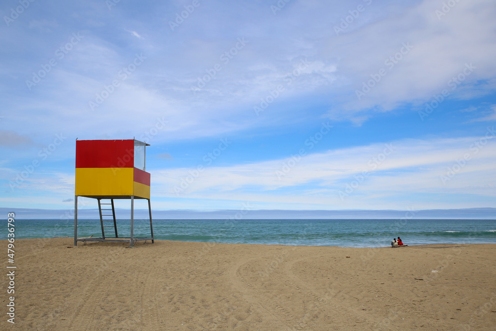 A surf life saving stand is a colourful focal point on a sandy beach. Two people sit on a log and enjoy the sea view on a warm summer day. Waipatiki Beach, North Island, New Zealand.
