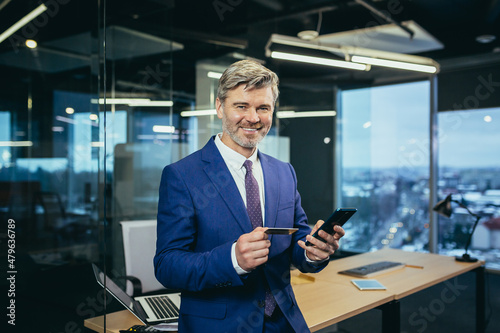 Experienced gray-haired businessman with a beard in a modern office, a man shopping online using a mobile application and phone © Liubomir