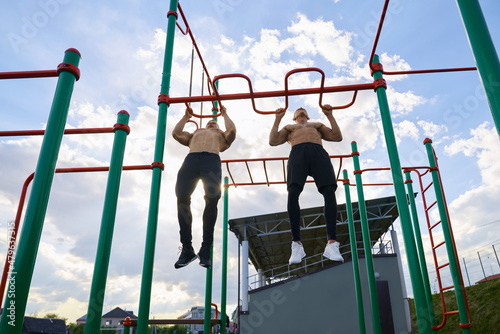 Low angle view of strong caucasian men doing exercises for biceps on sports ground. Two bodybuilders with bare torso pulling up on sport bars outdoors.
