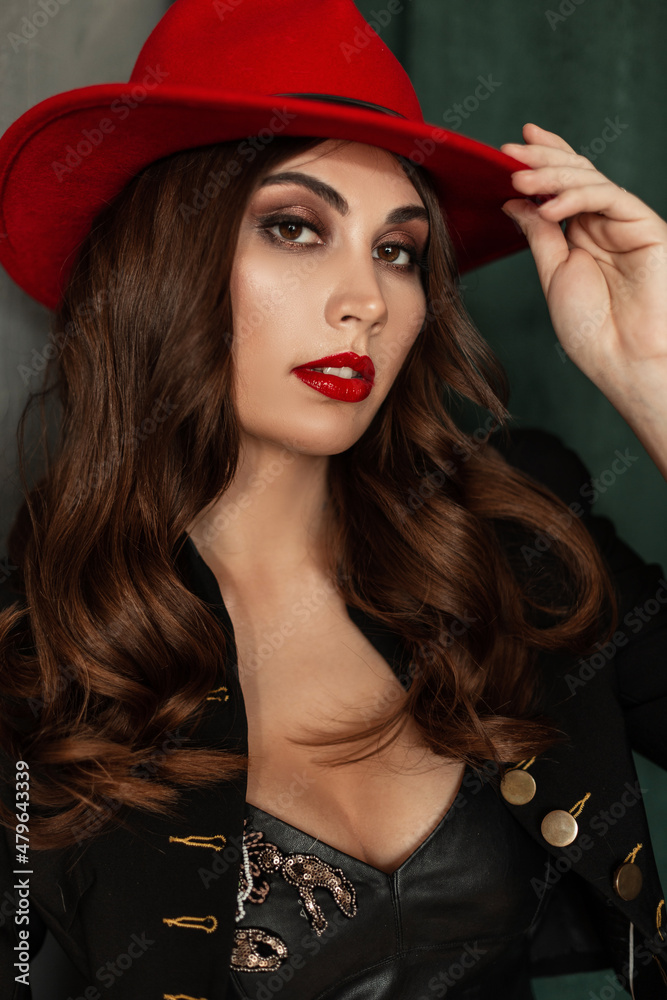Pretty sexy woman model with makeup, red lips and curly hairstyle in fashion elegant clothes look with blazer and red hat poses near vintage background Pretty sexy woman model with makeup, red lips and curly hairstyle in fashion elegant clothes look with blazer and red hat poses near vintage background