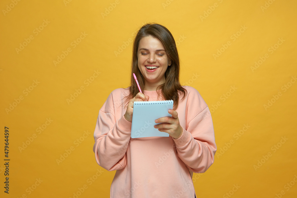 Laughing creative young girl writing in notebook, filling schedule, planning goals, create ideas on yellow background