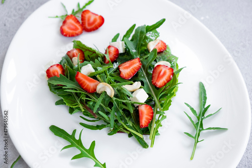 Romantic salad of strawberry with mix of herbs, arugula, spinach leaves, feta cheese and cashew nuts in heart shape on white plate. Romantic meal for St Valentine Day celebration.