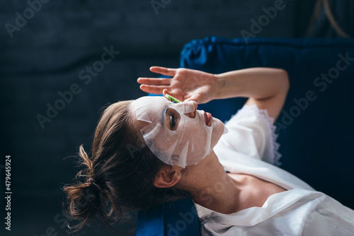 Woman lies on a couch with a sheet mask over her face and cucumber slices