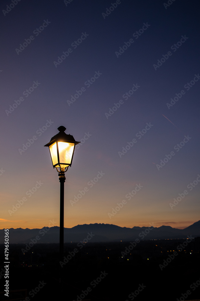 Moncalieri, Italy, beautiful sunset, lamppost, in the background mountains and houses, sky in shades of yellow and blue.