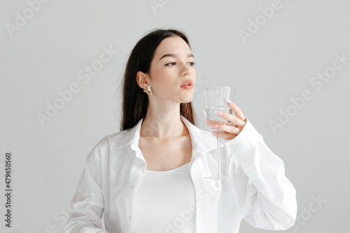 Studio portrait of young brunette woman in a white shirt and top, drinking glass of pure water, looking to the side, on white background. Purity concept