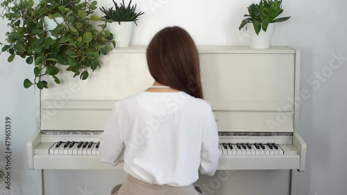 Back front view of unrecognizable young woman pianist playing on white classical piano in light room. Talented female musician student practicing in classroom during lesson. Shooting in slow motion