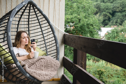 Young woman sitting in a hanging chair with a blanket and a cup of coffee on the balcony. © tumana_net