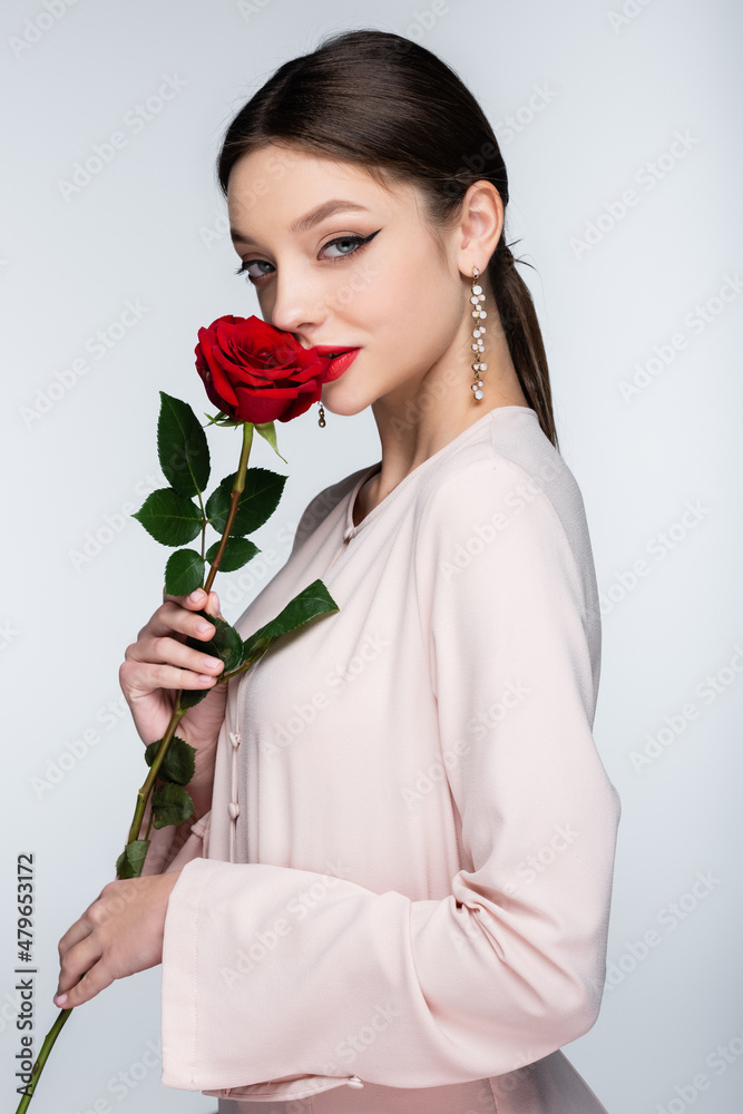 brunette woman in earrings and blouse smelling red rose isolated on grey
