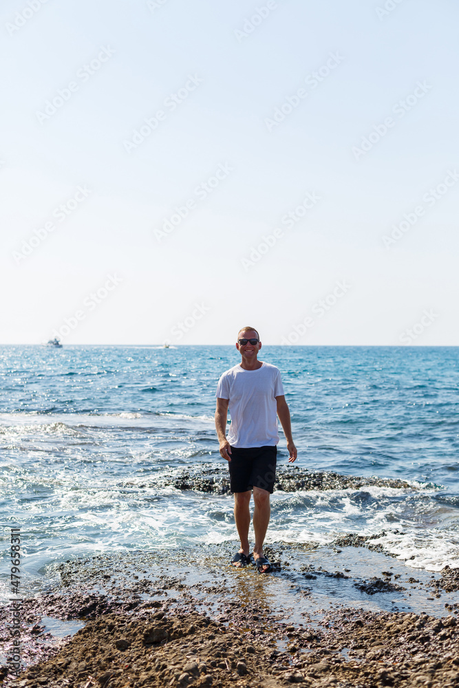 Young attractive man in sunglasses in a white t-shirt and shorts stands on the shore of the mediterranean sea