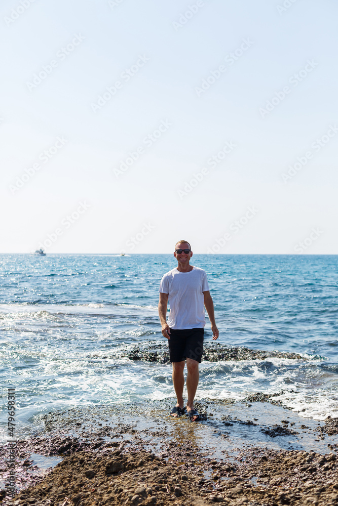 Young attractive man in sunglasses in a white t-shirt and shorts stands on the shore of the mediterranean sea Young attractive man in sunglasses in a white t-shirt and shorts stands on the shore of the mediterranean sea
