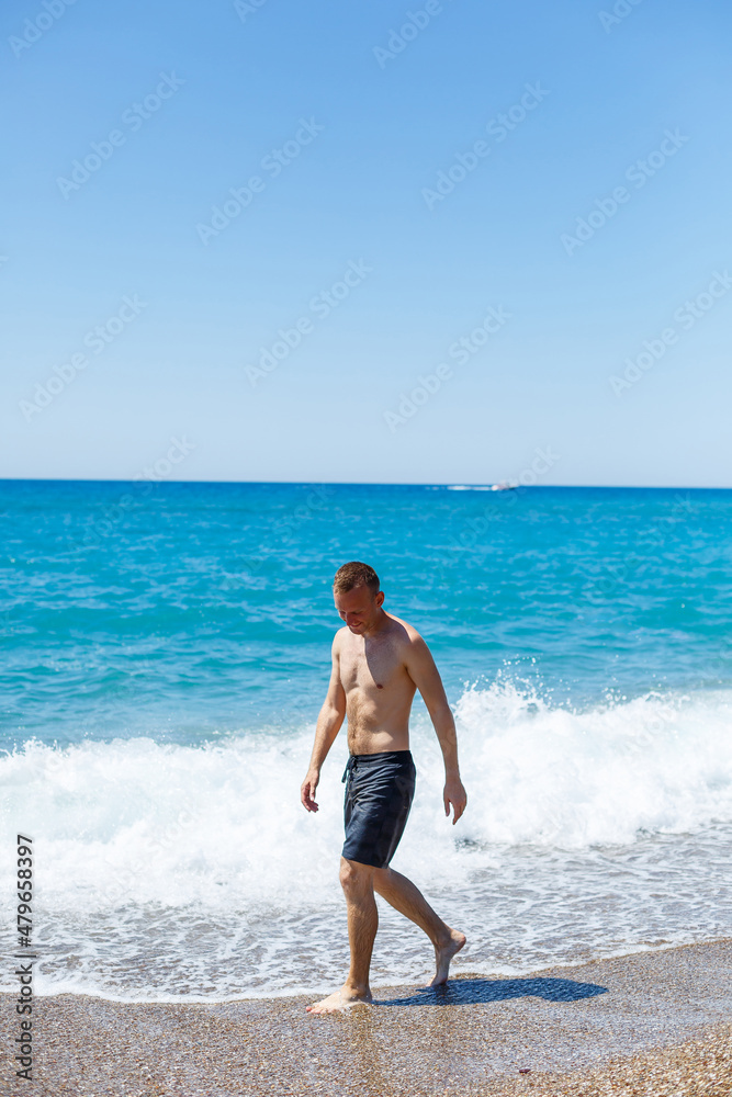 Attractive young man with a beautiful body in shorts walks along the sandy seashore and enjoying summer vacation Attractive young man with a beautiful body in shorts walks along the sandy seashore and enjoying summer vacation