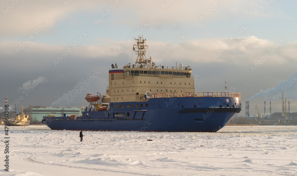 Fototapeta premium A fisherman catches fish on a frozen river and looks at the ships. Winter navigation on the river. Ice fishing.