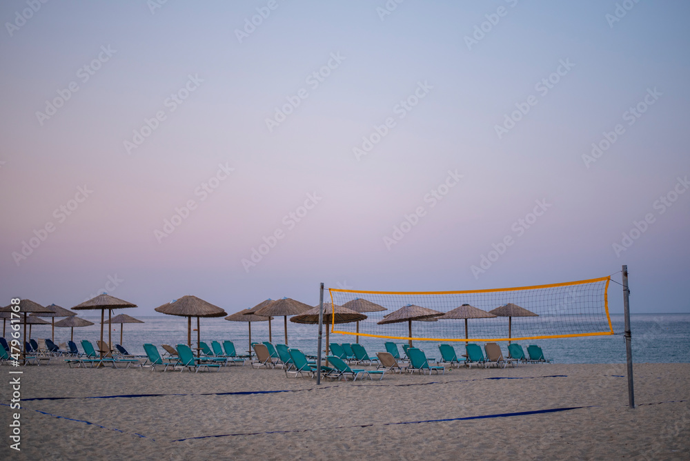 Fototapeta premium Beach Volleyball net on sandy beach early in the morning, with umbrellas and sunbeds, sea and blue sky in background.