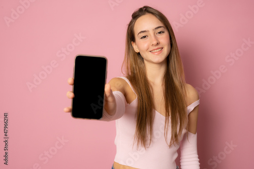 Close up portrait of young woman showing mobile screen over pink background.