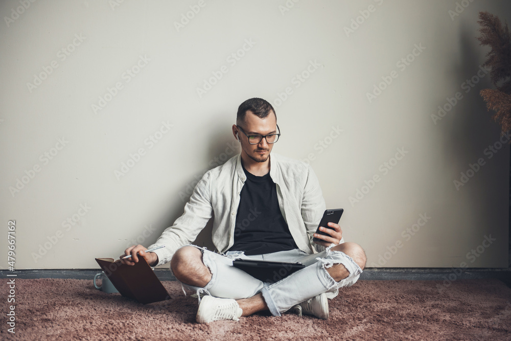 Front view of a caucasian man sitting on the floor looking at the smartphone while holding a book. Mobile communication. Online education. Front view of a caucasian man sitting on the floor looking at the smartphone while holding a book. Mobile communication. Online education.