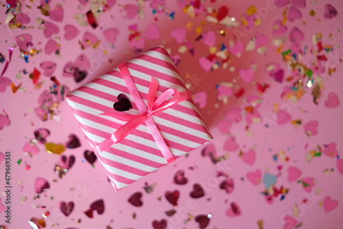 Foto Present in a striped paper wrapper with a pink ribbon on a background with a multicolored shiny confetti of hearts for Valentine's Day