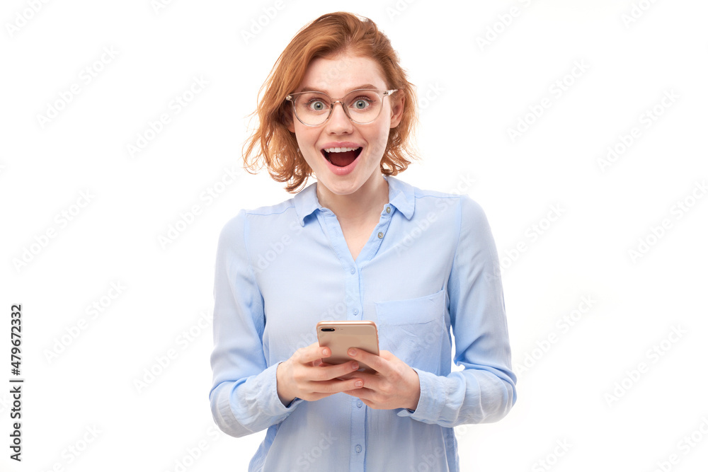Happy smiling redhead girl holding smartphone in hands in spectacles and business shirt isolated on white studio background, received a joyful message Happy smiling redhead girl holding smartphone in hands in spectacles and business shirt isolated on white studio background, received a joyful message
