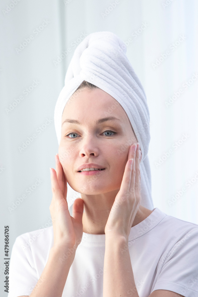 A beautiful middle-aged woman after a shower, in a white towel on her head, applies cream to her face, does spa procedures. Natural beauty of a woman. Vertical portrait A beautiful middle-aged woman after a shower, in a white towel on her head, applies cream to her face, does spa procedures. Natural beauty of a woman. Vertical portrait