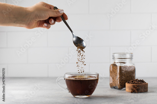 Woman stirs instant coffee in glass mug with boiled water on grey stone table photo