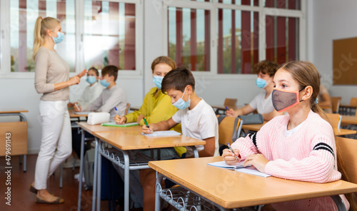 School girl in face mask sitting at desk in classroom on background with classmates and teacher