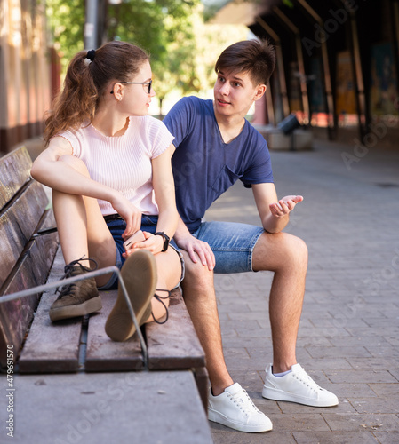 Smiling teenager boy talking with cute girl sitting on bench on city street on sunny summer day © JackF