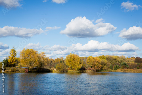 Autumn forest behind the lake. Sky with sun and white clouds. Red-green forest.