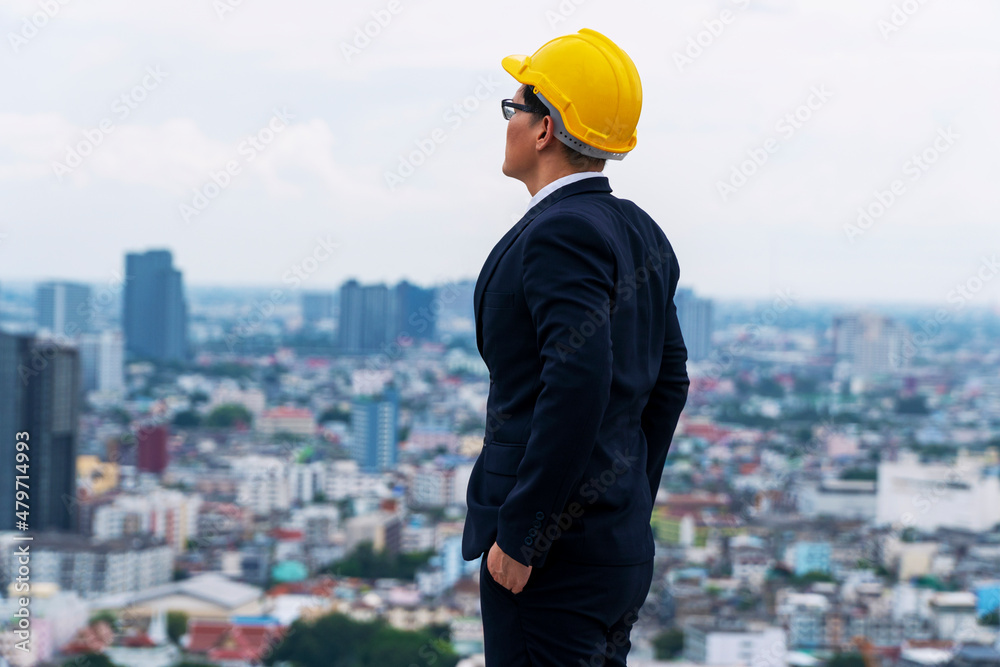 Confident architect asian businessman or formal wear yellow safety hardhat thinking plant work standing top builder with skyscraper city view background. Manger engineer with helmet looking building. Confident architect asian businessman or formal wear yellow safety hardhat thinking plant work standing top builder with skyscraper city view background. Manger engineer with helmet looking building.
