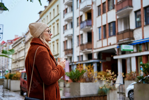 Young woman in eyeglasses wearing coat walking at city street with cup of coffee takeaway