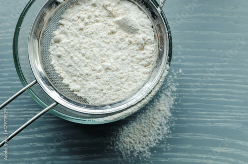 Fotografie Wheat flour is sown through a sieve into a glass bowl through a metal sieve, on a wooden table