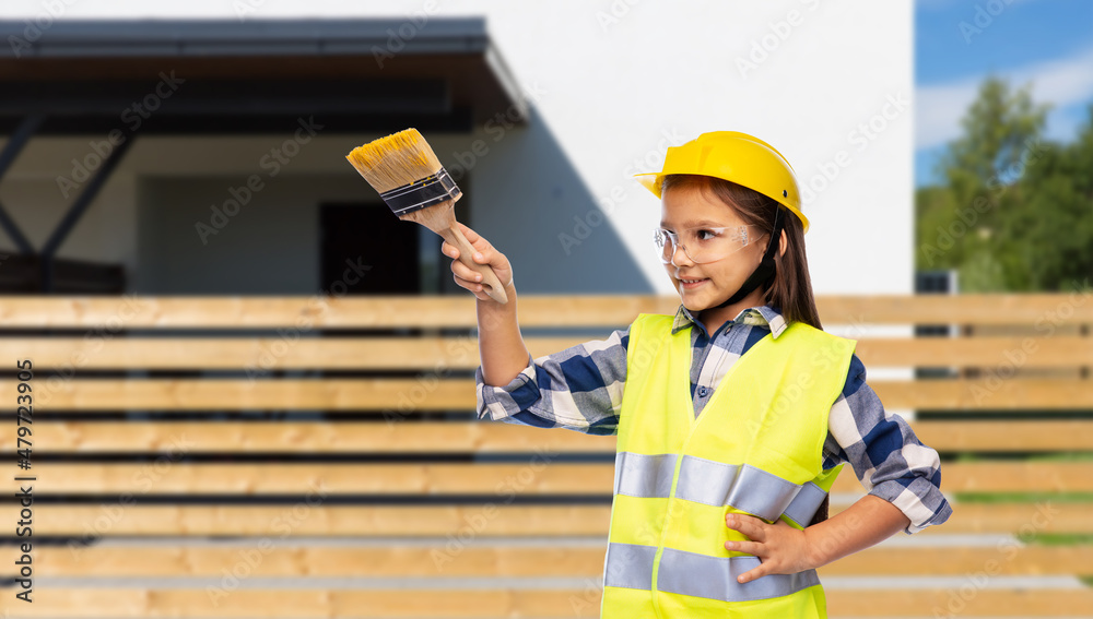 building, construction and profession concept - smiling little girl in protective helmet and safety vest with paint brush over house background building, construction and profession concept - smiling little girl in protective helmet and safety vest with paint brush over house background