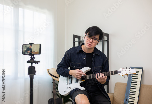 Young asian musician teaching the rock guitar class online live streaming on smartphone with student. Korean man playing electric guitar singing a song and record by cell phone shaed online social photo