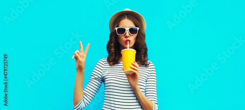 Portrait of stylish young woman drinking a fresh juice wearing a summer straw hat and striped t-shirt on blue background